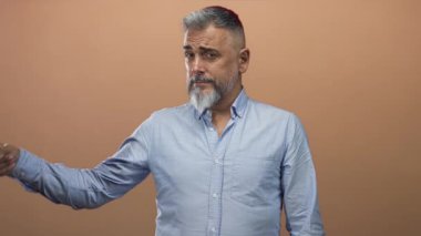 Stüdyoda şeftali arkaplan ve açık mavi gömlekli, gri sakallı ve kaşlarını kaldırmış bir adam, alaycı mizah şüpheciliği.