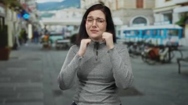 Kumral saçlı bir kadın, şehir manzarası bulanık, gözlüklü ve gri kazaklı, hayal kırıklığı ve tefekkür sergileyen, açık bir sokakta duygusal ifadeler sergiliyor..