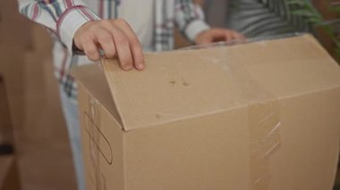 Adam, içinde bina içi olan karton bir kutuyu parlak ekose gömlek giyerek açıyor; taze bir başlangıç heyecanı.