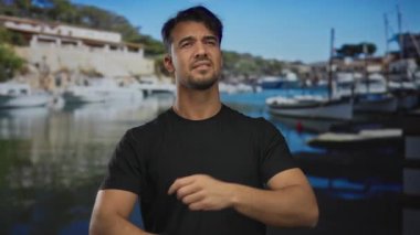 Düşünceli bir suratı olan genç İspanyol bir adam tekneleri ve sakin bir açık hava ortamı olan manzaralı bir sahil limanında duruyor..