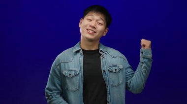 Kot giyen genç adam, izole edilmiş mavi arka plana karşı özgüvenle el sallıyor.