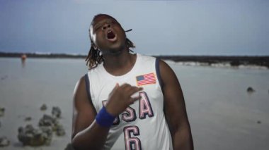 Adamım, genç Afro-Amerikalı, ağzı açık ve kafası eğilmiş, plaj şeridinde bayrak yaması olan ABD forması giymiş, sığ su ve kayalar görülebilir..