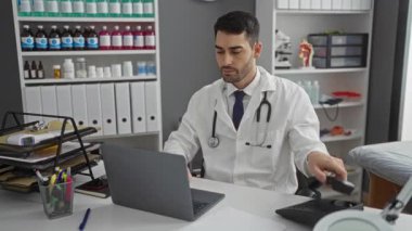 Beyaz önlüklü genç adam telefonla konuşuyor ve bir klinikte dizüstü bilgisayar kullanıyor. Arka planda raflar dolusu malzeme var..