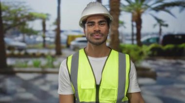 Adam, palmiye ağaçları ve park halindeki arabalarla kaplı kentsel caddede yansıtıcı yelek giyerken beyaz kaskı ayarlar; güvenlik garantisi.