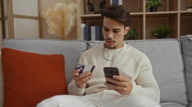 Genç adam, modern bir oturma odasındaki rahat bir koltukta otururken elinde kredi kartı ve akıllı telefon tutuyor. Şık bir apartmanın internet aktivitelerini ve finansal planlarını yansıtıyor..
