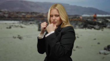 Kumsalda takım elbiseli bir kadın, güçlendirme konsepti için sahilin manzaralı arka planına karşı yumruklarıyla poz veriyor.