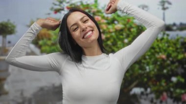 Açık havada uzanan ve el kol hareketleri yapan bir kadın, yeşil yaprakların arasında gülümseyen, güneşli bir günde beyaz uzun kollu bluz giyen, rahatlama ve neşe önerisi sunan..