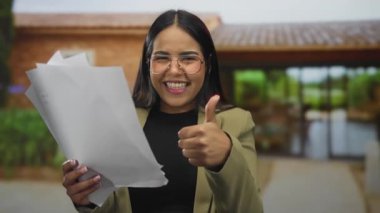 Dışarıda elinde başparmağıyla belgelerle gülümseyen, gözlük takan, spor ceket giyen, neşe ve başarı tasvir eden bir kadın..