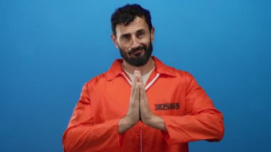 Turuncu hapishane tulumu giyen İspanyol sakallı adam, sakin ve içsel bir duayı gösteren mavi bir arkaplanda poz veriyor..