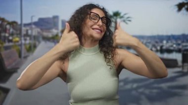 Kıvırcık saçlı ve gözlüklü kadın güneşli sahil caddesinde çifte onay veriyor. İyimserlik teşvik..