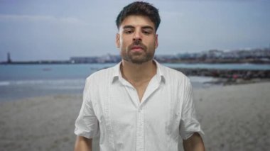 Beyaz gömlekli adam mavi gökyüzünün altında rüzgar saçı ve güneş parıltısıyla deniz kenarını çimdikler; iğrenme.