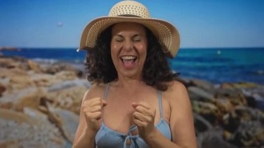 Güneşli plaj gününün tadını çıkaran bir kadın bikini ve şapka giyiyor açık havada deniz kenarında gülümsüyor ve neşe saçıyor..