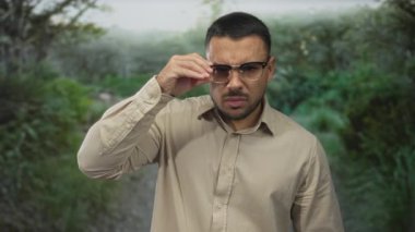 Gözlüklü ve bej gömlekli adam yeşil yapraklar arasında orman yolu boyunca parmağını işaret ediyor; iddialı yüzleşme.