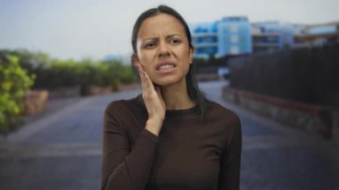 Sessiz bir sokakta acı içinde çene tutan İspanyol bir kadın. Yeşillik ve binalar yumuşak odak noktasında. Kentsel bir ortamda rahatsızlık ve endişeyi dile getiriyor..