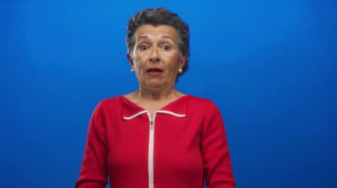 Kırmızı kazaklı, gri saçlı yaşlı bir kadın canlı mavi bir arkaplan karşısında şok olmuş görünüyor. Bu görüntüde canlı bir şaşkınlık ve duyguyu ifade ediyor..