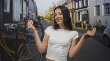 Sokakta avuç içi omuz silken, beyaz hasat bluzu giyen ve şehir evleri ve park edilmiş bisikletlerin arasında gülümseyen bir kadın..