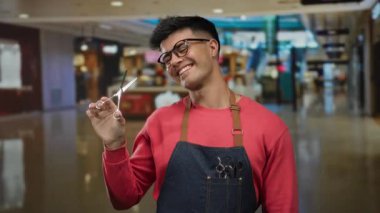 Önlüklü, elinde makasla alışveriş merkezindeki bir dükkanda güvenle gülümseyen ve işe hazır berberi canlandıran İspanyol asıllı genç bir adam..