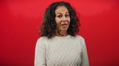 Kıvırcık saçlı olgun İspanyol bir kadın canlı kırmızı bir arka planda jestler yapıyor. Zarafeti vücut dili ile anlamlı iletişimi birleştiriyor..