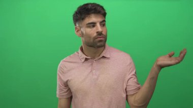 Açık pembe polo giyen İspanyol adam, omuz silkme ifadesi gösteren açık el hareketleriyle yeşil stüdyoda el ele tutuşuyor..