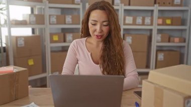 Bir kadın depolama binasındaki paket kutularının arasında bilgisayardan bakıp gülümsüyor. Küçük işletme keyfi..