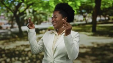 Açık hava parkında gülümseyen bir kadın iki eli havada, beyaz ceketli ve yeşil ağaçlarla çevrili neşeli bir jest yapıyor..