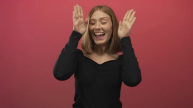 Siyah tişörtlü, sarı saçlı genç bir kadın, izole edilmiş kırmızı bir duvara karşı duruyor. Yüzünü kapayarak, ilginç ve dinamik bir görüntü yaratıyor..