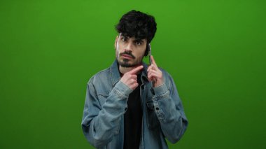 Genç İspanyol bir adam yeşil arka planda bir telefonla konuşurken sessizlik için el kol hareketi yapıyor, kot ceket giyiyor ve kameraya dönük çeşitli duygular ifade ediyor..