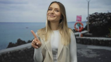 Kadın gülümsüyor ve parmaklarıyla parmakla işaret ediyor. Deniz kenarında gezinirken, arka planda okyanus var..