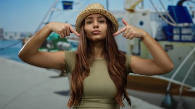 Şapkalı genç bir kadın arka planda teknelerle limanı işaret ediyor açık mavi gökyüzünün altında canlı bir deniz manzarası sergiliyor kendine güvenen bir ifadeyle..