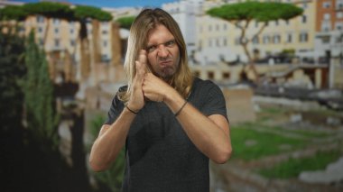 Sarışın adam, arka planda antik Roma harabeleri olan bir binanın içinde el hareketini engellemek için avucunun içinde yumruk tutuyor..