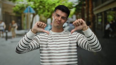 Çizgili gömlekli genç adam şehir caddesinde bulanık ağaçlar ve insanlarla başparmak kaldırıyor. Açık hava ortamını tasvip etmiyor veya hayal kırıklığına uğratmıyor..