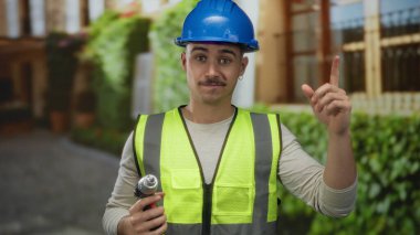 Yansıtıcı yelek ve kasketli genç İspanyol bir adam şehir caddesinde bir matkap tutuyor, açık hava ortamında bir fikir belirtiyor, inşaat çalışmalarını sergiliyor..