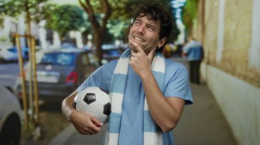 Hispanik bir adam sokakta, elinde düşünceli bir ifadeyle futbol topu tutuyor, şehir sporu hevesini somutlaştırıyor..