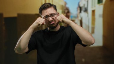 Sakallı ve gözlüklü genç bir adam, şehir sokaklarında ağlıyormuş gibi yapıyor oyuncu ifadeler ve komik duygular sergiliyor..