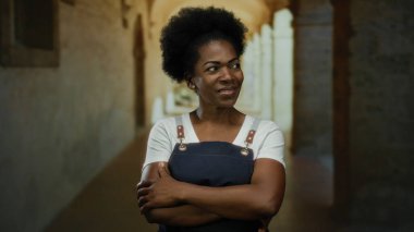 Önlük takmış, kollarını kavuşturmuş kendinden emin bir kadın kemerli koridorları olan eski bir üniversitede kapalı bir yerde duruyor ve gururunu ve güvenini dile getiriyor..