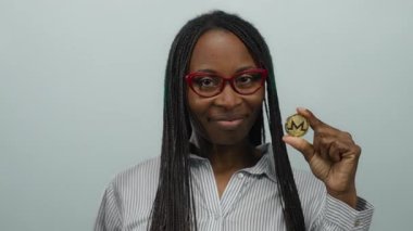 Gözlüklü Afrikalı Amerikalı bir kadın beyaz bir duvara monero bozuk para tutuyordu. Sanal para konsepti sergiliyordu. Kendine güvenen bir ifadesi ve profesyonel kıyafeti vardı..