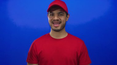 Kırmızı tişört giyen İspanyol kurye stüdyoda açık ağızlı ve parlak mavi arka planlı başparmaklar..