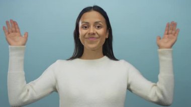 Beyaz kazaklı genç bir kadın ellerini kaldırmış mavi arka plan duvarına gülümsüyor ve selamlıyor..