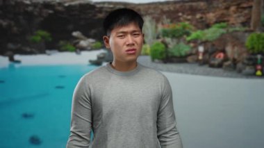 Serin bir havuzun yanında duran genç adam açık havada, doğa ve yeşilliğin ortasında rahat bir atmosfer sergiliyor.