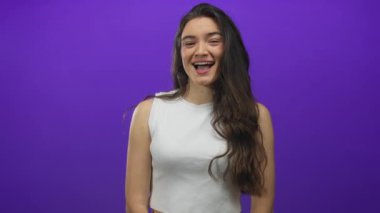 Latin kökenli genç bir kadın mor bir stüdyoda uzun siyah saçlı, hafif ışıklandırma altında göz kırpıyor..