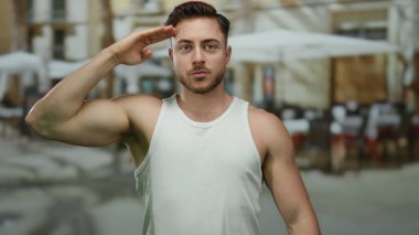 Şehir restoranının terasında oturan sakallı genç adam arka planda bir sokak dekorunu gösteriyor..