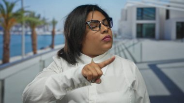 Deniz kenarındaki gezinti alanında el kol hareketi yapan genç Latin bir kadın, gözlük ve beyaz gömlek takıyor, palmiye ağaçları ve binaların arasında tavır sergiliyor..