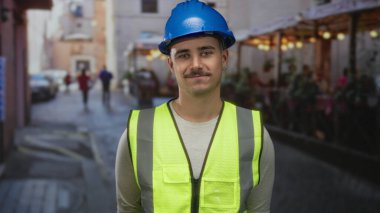 Yansıtıcı yelek giyen genç İspanyol bir adam bir restoran ve kahve dükkanının terasında duruyor, iş ve rahatlama karışımı bir şey taşıyor..