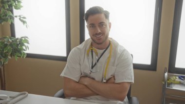 Doktor kıyafeti ve steteskop giyen sakallı genç adam kollarını kavuşturup klinik ortamında profesyonel bir sağlık hizmeti sunuyor..