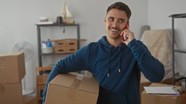 Genç adam yeni bir apartman dairesinde karton bir kutu tutarken akıllı telefondan konuşuyor. Etrafı eşyalarla çevrili, hareket sürecini sembolize ediyor..