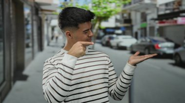 Şehir sokağındaki palmiyeyi gösteren genç adam, çizgili gömlek giyiyor, kendine güvenen ve çekici bir pozla açık şehir atmosferini yakalıyor..
