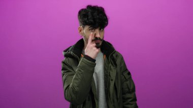 Yeşil ceketli genç İspanyol adam, tetikte olduğunu ve dikkat ve dikkat gösterdiğini belirten pembe bir arka planda parmağı ile ihtiyatlı bir jest yapıyor..