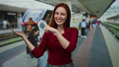 Tren istasyonunda gülümseyen, kırmızı giyen, elini gösteren, ayakta duran, dışarıda duran, kızıl saçlı, güzel, yetişkin, çekici, gösterişli seyahat ya da ulaşım sahnesi olan bir kadın..