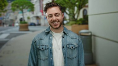 Sakallı genç adam güpegündüz şehir sokaklarında kot ceket giyerek ağaçların ve binaların arasında gülümsüyor..