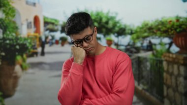 Gözlüklü ve kırmızı gömlekli genç bir adam şehir sokağında dikiliyor, yeşil ve taştan duvarları olan bulanık şehir zeminine karşı düşünceli görünüyor..